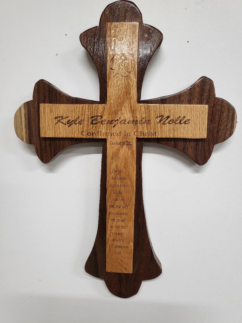A cross made of wood and words carved on it and hung on a white wall
